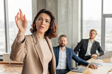 Aufkleber - A woman in a beige blazer stands in front of a conference table, leading a business meeting with two colleagues seated behind her. She is gesturing with her hand, engaging in a presentation