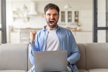 Poster - A man with a beard sits on a beige couch in a living room and excitedly celebrates with a raised fist, looking directly at the camera while using a laptop.