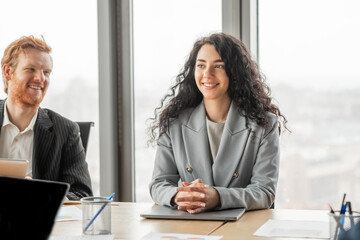 Poster - A businesswoman with long curly hair smiles and sits across from a male colleague at a conference table in a modern office setting.