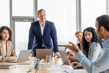 Aufkleber - A group of business professionals are gathered around a table, discussing ideas. One man is standing, looking at the group while another man gestures, engaged in the conversation