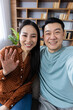 © Liubomir - Asian couple sitting in living room making video call using phone, smiling and waving at camera. Cozy home environment with bookshelf in background