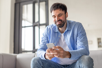 Poster - A man with a beard sits on a couch in a light-colored room, smiling as he uses his phone. He is wearing a blue button-down shirt and blue jeans.