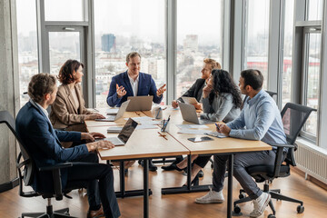 Poster - A group of business professionals are gathered around a large conference table in a modern office, engaging in a discussion. Laptops and documents are spread out on the table