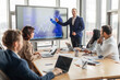 © Prostock-studio - A business professional stands near a large digital screen showing an upward trending line graph. The presenter gestures towards the screen as they explain the chart to a group of colleagues
