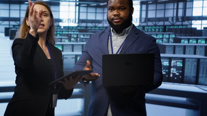 Poster - Teamworking colleagues doing checkup, inputting data on laptop of needed server hub fixes. Coworkers working together in data center housing advanced storage infrastructure, camera A