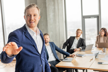 Aufkleber - A businessman in a blue suit stands and addresses his colleagues who are sitting at a conference table during a business presentation.
