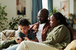 © pressmaster - Happy young African American couple looking at their cute little son scrolling in smartphone while spending weekend at home
