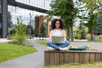 African American woman using laptop in outdoor park setting near modern office building. Woman smiling, sitting on bench with phone and coffee, enjoying work in nature and city scene.