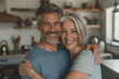 © GraysonStock - A bearded elderly man and his gray-haired wife hugging and smiling, standing in the kitchen of their home.