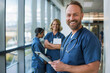 © GraysonStock - A middle-aged male nurse in a blue uniform smiling and holding a tablet looking straight ahead.