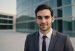© SerPak - A young businessman in a suit stands outdoors confidently near a glass-walled office building, exuding professionalism and ambition with a modern architectural backdrop.