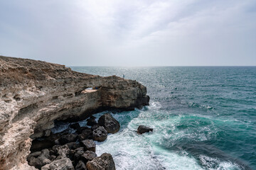 Wall Mural - The ocean is calm and the sky is cloudy. The water is a deep blue color. The rocks are jagged and the waves are small.