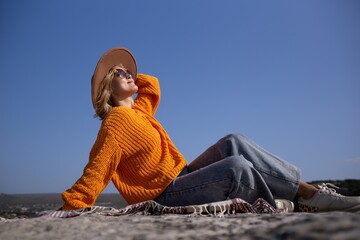 Wall Mural - A woman in an orange sweater and blue jeans is sitting on a blanket on a rock