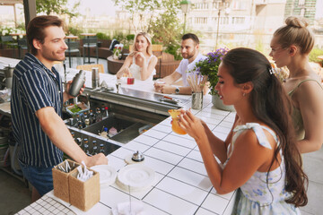 Wall Mural - Young people, friends meeting in modern bar, open air restaurant, enjoying cocktails and conversation with male bartender. Concept of friendship, party, leisure, celebration, relaxation