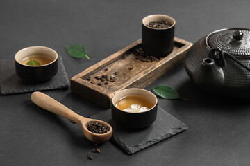 Sticker - Teapot with cups, wooden spoon of dry tea and leaves on black background