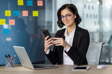 Wall Mural - Confident businesswoman in glasses using phone in modern office. Smiling professional working on laptop surrounded by colorful sticky notes, showcasing productivity and modern business environment.