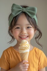 Smiling young girl with green bow and yellow shirt holding and enjoying a vanilla ice cream cone.