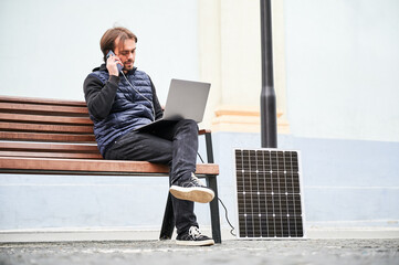 Man sits on wooden bench, working on laptop and smartphone, both connected to solar panel. Urban setting and casual attire highlight modern, sustainable, mobile workspace.