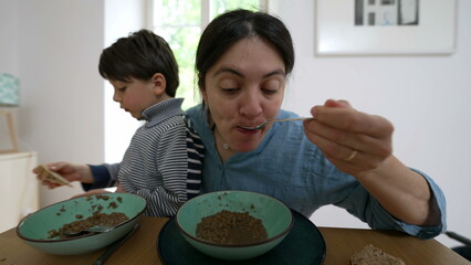 Mother multitasking by feeding her young son and eating her meal at the dining table. moment of family bonding and the daily routine of caring for a child during mealtime