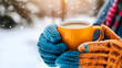 © Tanya - Hands in knitted mittens holding yellow ceramic coffee cup in snowy forest. Christmas, wintertime concept. Copy space.