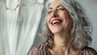 © GoodandEvil - Woman with gray hair laughing near white curtains happily.