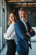 © GraysonStock - A mature businessman and a young businesswoman stand with arms crossed side by side, looking at the camera smiling.