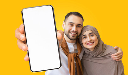 Fototapete - A young Muslim couple smiles at the camera while holding a smartphone with a blank screen. They are standing in front of a bright yellow background. The woman is wearing a hijab