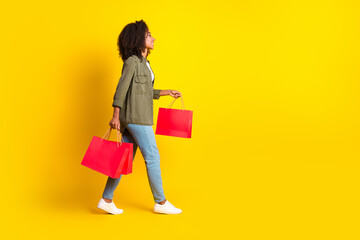 Full size side profile photo of nice girl wear khaki shirt walk with shopping bags to empty space isolated on yellow color background