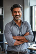 © GraysonStock - A Hindu businessman standing happily in an office crossing his arms, dressed casually in a shirt.