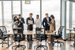 © Prostock-studio - A diverse group of business professionals stand around a conference table in a modern office space. They are engaged in a lively discussion