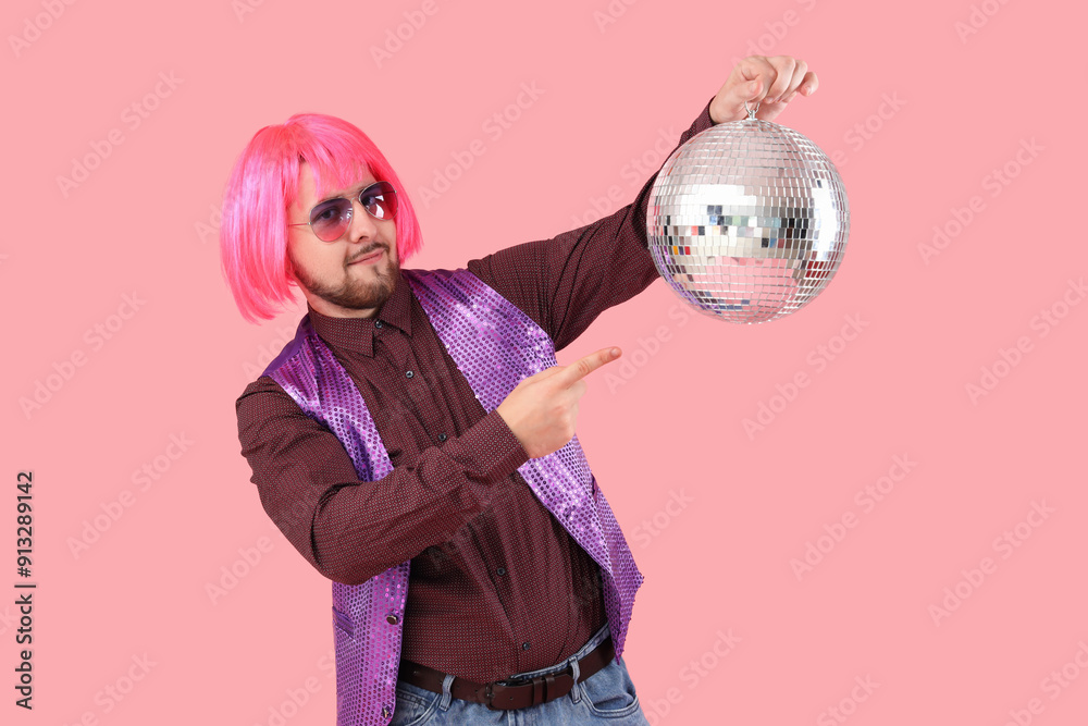 Handsome young man in wig pointing at disco ball on pink background