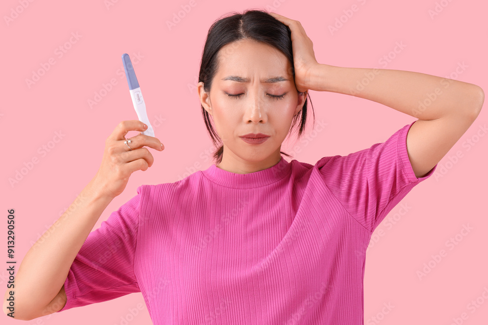 Upset Asian woman with pregnancy test on pink background