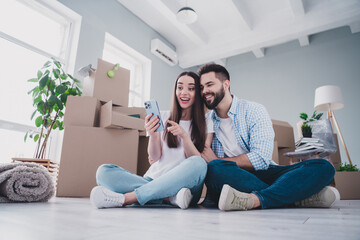 Wall Mural - Full body portrait of two young people use phone sit floor relocating moving box package new apartment indoors