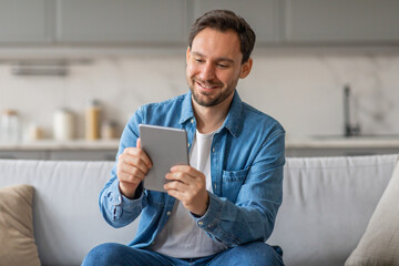 Poster - A man in a blue shirt is sitting on a white couch in a living room. He is smiling as he looks at a tablet in his hands.