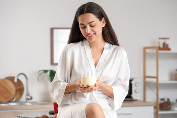 Poster - Pretty young woman with cup of hot coffee sitting on kitchen counter at home