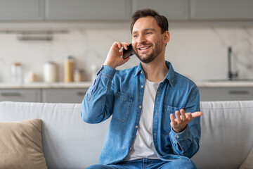 Poster - A man with a beard is sitting on a white couch in his home while talking on his phone. He is wearing a blue denim shirt and jeans. He is smiling and gesturing with his hand as he speaks.