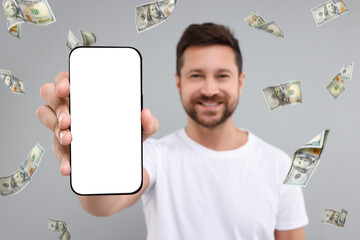 Wall Mural - Happy man showing mobile phone with empty screen under money rain on grey background, selective focus