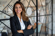 © GraysonStock - A smiling young Latina woman, dressed in a black suit, standing with her arms crossed, a female manager of a finance company.