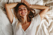 © BetterPhoto - A woman relaxing in bed with a smile, wearing a white shirt, representing comfort and peace.