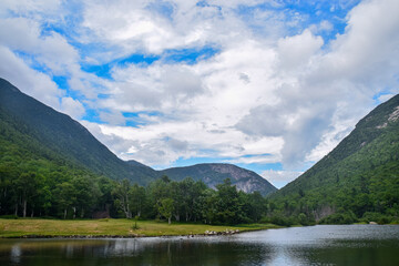 Wall Mural - Serene lake surrounded by lush green mountains and a blue sky.