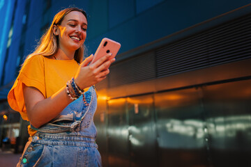 Copy space. One happy young girl using her smartphone to browse the internet while walking on the street at night. Female watching the social media with her cellphone. Girl typing on a mobile phone