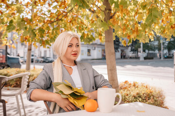 Wall Mural - A blonde woman sits at a table with a cup of coffee and a stack of papers. She is wearing a gray jacket and a necklace. The scene is set in a park with trees and benches.