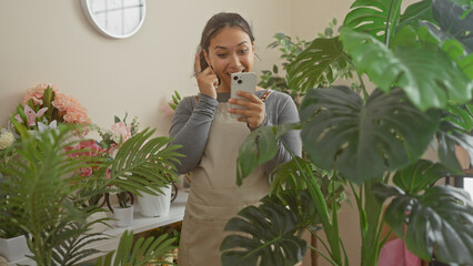 Sticker - A young hispanic woman in an apron smiles while using a smartphone among vibrant indoor plants in a flower shop.