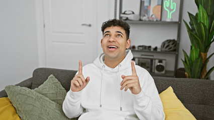 Wall Mural - A smiling young man with curly hair sitting on a sofa indoors, pointing upwards with both hands, giving off an excited and optimistic vibe.