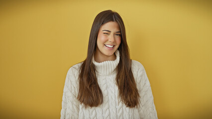 Poster - A cheerful young hispanic woman in a white sweater poses against an isolated yellow background, exuding joy with a playful wink.