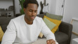 © Krakenimages.com - A thoughtful young african american man wearing a white sweater, sitting indoors in a well-decorated living room.