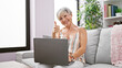 © Krakenimages.com - A smiling mature woman with grey hair gives a thumbs-up while using a laptop on a sofa indoors.