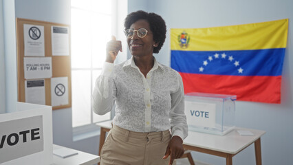 Middle-aged woman with curly hair giving a thumbs up in an indoor venezuelan electoral room with a flag and vote signs in the background