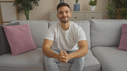 Canvas Print - Handsome young hispanic man with a beard sitting in the living room of his home, looking relaxed and cheerful while wearing a white t-shirt and jeans.