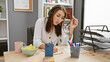 © Krakenimages.com - A concentrated brunette woman with headphones working in a modern office, writing notes and surrounded by technology and snacks.